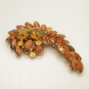 May include: A gold-tone brooch featuring a cluster of amber-colored faceted crystals in various shapes, including round, oval, and marquise cuts, arranged in a curved, leaf-like pattern with prong settings securing each individual stone.