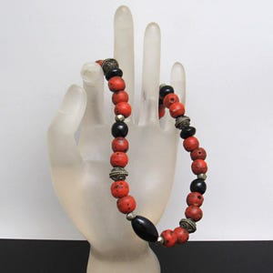 May include: A bracelet with red and black beads, interspersed with silver-coloured metal elements. The bracelet is displayed on a clear, hand-shaped form. The beads are round and have a textured finish, creating a unique accessory.