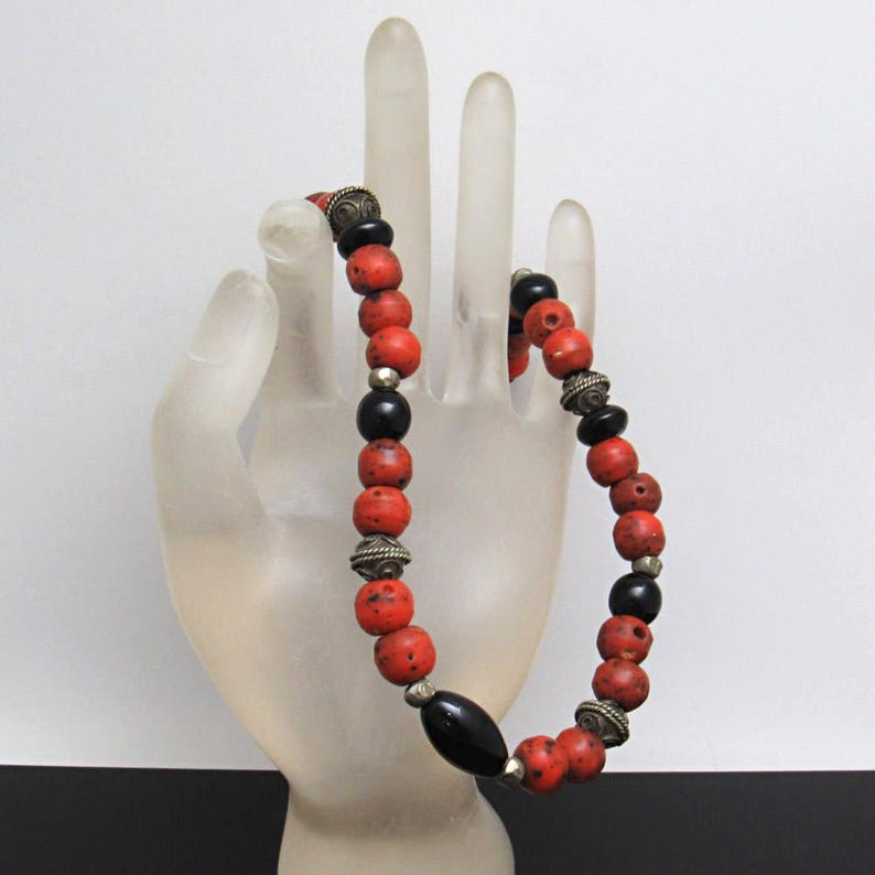 May include: A bracelet with red and black beads, interspersed with silver-coloured metal elements. The bracelet is displayed on a clear, hand-shaped form. The beads are round and have a textured finish, creating a unique accessory.