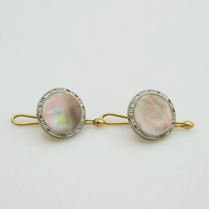 May include: A pair of cufflinks featuring circular mother-of-pearl inlays with iridescent pink and white tones. Each inlay is set within a silver-toned frame with a textured edge, connected by a gold-toned bar and a curved toggle mechanism.