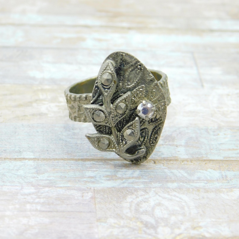 May include: A silver-toned ring with an oval centerpiece featuring a leaf design and small, round embellishments. A single, clear gemstone is set in the center. The band has a textured, bark-like appearance.