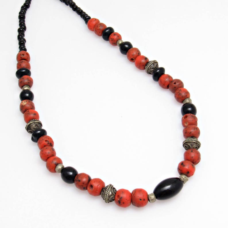 May include: A necklace with red, black, and silver beads. The beads are various shapes, including round and oval. The necklace is strung on a black cord. The red beads have a speckled appearance, and the silver beads have a decorative design.