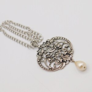 Puede incluir: Un collar de plata con un delicado colgante adornado con una perla. El colgante est&aacute; hecho de filigrana intrincada y tiene una forma circular.