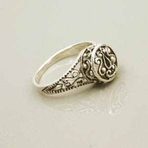 Pu&ograve; includere: Un anello in metallo color argento caratterizzato da un intricato design in filigrana traforata sulla fascia e un elemento centrale circolare rialzato con motivi decorativi a volute e piccoli accenti sferici.