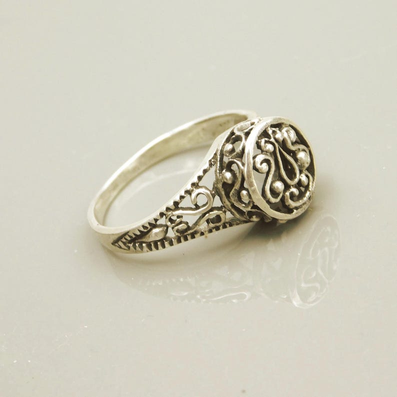 Pu&ograve; includere: Un anello in metallo color argento caratterizzato da un intricato design in filigrana traforata sulla fascia e un elemento centrale circolare rialzato con motivi decorativi a volute e piccoli accenti sferici.