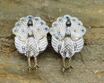 Op de afbeelding: Een paar witte emaille pauwenbroches met gouden accenten en blauwe ogen. De pauwen kijken in tegengestelde richtingen.