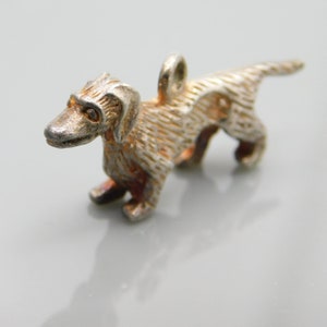 May include: A silver charm in the shape of a dachshund dog. The charm is detailed with textured fur and has a loop for attaching to a bracelet or necklace.
