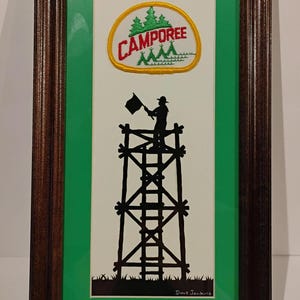 May include: A framed artwork featuring a silhouette of a person standing on a tall wooden tower, holding a flag. The tower is made of wooden beams and has a ladder leading up to the top. The artwork is set against a green background and includes a patch with the text "CAMPOREE" in red and yellow.