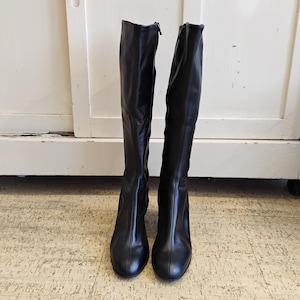 Könnte beinhalten: Ein Paar schwarze kniehohe Stiefel mit runder Spitze und kleinem Absatz. Die Stiefel haben einen Reißverschluss an der Seite und sind aus glattem, glänzendem Material gefertigt. Die Stiefel stehen auf einem gemusterten Boden.