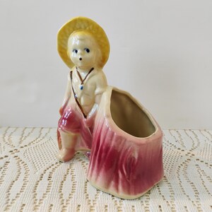 Può includere: Fioriera vintage in ceramica a forma di bambino con cappello giallo e abito rosa e bianco. La fioriera ha una grande apertura per fiori o altri oggetti decorativi. La statuina ha gli occhi blu e una collana.