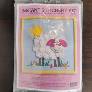 May include: An Instant Stitchery Kit in a clear plastic bag featuring a lamb design. The package shows a finished 12 x 12 inch project with a white loomed wool lamb, a yellow sun, and colorful flowers on a light blue background with a yellow frame.