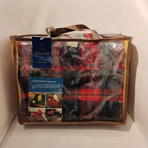 Puede incluir: Una bata Pendleton en una bolsa de plástico transparente con un asa marrón. La bata tiene un estampado de cuadros rojos y negros. La bolsa tiene dos etiquetas con el logotipo de Pendleton e información del producto.