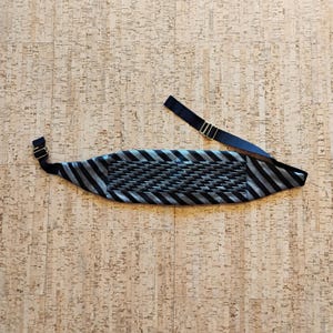 Puede incluir: Un fajín a rayas negras y plateadas con una correa negra ajustable y herrajes dorados. El fajín tiene una sección central tejida y está diseñado para usarse alrededor de la cintura.