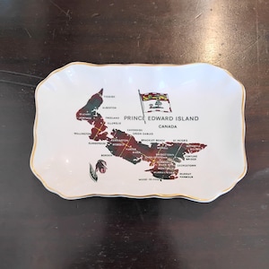 May include: A rectangular white ceramic tray with scalloped edges and a gold-trimmed rim. The centre features a map of Prince Edward Island, Canada, filled with a red and green tartan pattern, the provincial flag, and a pink lady slipper flower.