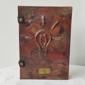 May include: A rectangular copper journal cover featuring a raised embossed lightbulb design in the center. The metal surface displays a mottled patina with brown and reddish tones. Two black leather straps with brass rivets secure the side.