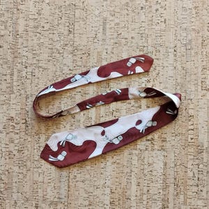 Puede incluir: Una corbata de color granate y crema con un patrón repetitivo de formas abstractas. La corbata está hecha de un material suave y sedoso y se muestra sobre una superficie texturizada. El diseño de la corbata presenta una combinación de colores granate y crema.