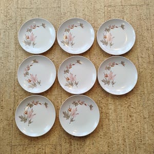 Op de afbeelding: Set van acht witte keramische borden met een bladerenmotief. Elk bord heeft een gebogen rangschikking van roze, bruine en gouden bladeren. De borden zijn gerangschikt op een getextureerd, lichtbruin oppervlak.