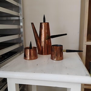 May include: A copper tea set with a teapot, a small pot, and a cup. The teapot has a long spout and a black handle. The small pot and cup have black handles. The set is on a white table.