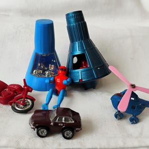 May include: A collection of small plastic toys including a red motorcycle, a maroon car, a blue helicopter with pink blades, a red and blue action figure, and two blue space capsule models, one of which has a clear window showing interior details.