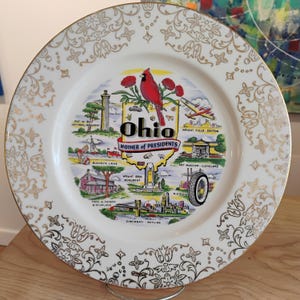 Puede incluir: Un plato decorativo blanco con un borde floral dorado y un diseño central con monumentos de Ohio. El diseño incluye un cardenal rojo, la palabra "Ohio" y el texto "Mother of Presidents".