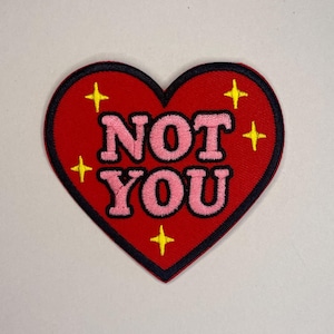 Könnte beinhalten: Ein roter herzförmiger Aufnäher mit den Worten "NOT YOU" in Rosa, schwarz umrandet. Der Aufnäher hat gelbe Sternakzente und einen schwarzen Rand. Dieser Aufnäher ist für Jacken, Taschen oder andere Stoffartikel geeignet.