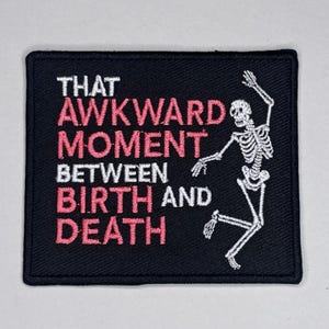 Könnte beinhalten: Schwarzer quadratischer Aufnäher mit dem Text "THAT AWKWARD MOMENT BETWEEN BIRTH AND DEATH" in Rosa und Weiß. Auf der rechten Seite des Aufnähers tanzt ein weißes Skelett.