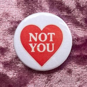 May include: A round white button with a red heart design. The heart contains the words "NOT YOU" in white capital letters. The button is set against a pink, textured background.
