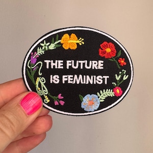 Könnte beinhalten: Ein ovaler, schwarz bestickter Aufnäher mit dem weißen Text THE FUTURE IS FEMINIST in der Mitte. Das Design ist von bunten gestickten Blumen und grünem Blattwerk umgeben, darunter gelbe, rote, rosa und hellblaue Blüten.