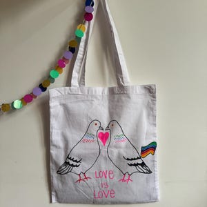 May include: A white cotton tote bag featuring a black line drawing of two pigeons facing each other with a bright pink heart between them. The design includes the text "LOVE IS LOVE" in pink and a small rainbow flag attached to one pigeon.