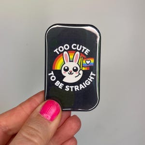 Könnte beinhalten: Schwarze rechteckige Anstecknadel mit abgerundeten Ecken. Die Anstecknadel zeigt ein weißes Kaninchen mit einem Regenbogen, einem Herzen und dem Text "TOO CUTE TO BE STRAIGHT" in Weiß. Das Kaninchen hält eine Flagge mit einem Herzen.