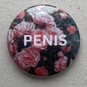 May include: A round button with a floral design featuring pink roses and the word "PENIS" in white block letters. The background is dark, creating a contrast with the bright flowers and text.