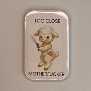 May include: A rectangular button featuring a cartoon lamb holding a knife. The lamb has a yellow collar and a flower on its side. The text "TOO CLOSE" is above the lamb, and "MOTHERFUCKER" is below.