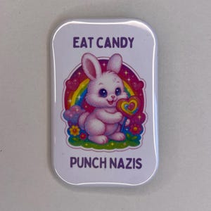 Könnte beinhalten: Ein rechteckiger weißer Button mit dem Text "EAT CANDY" und "PUNCH NAZIS" in Lila. Der Button zeigt ein Cartoon-Kaninchen, das einen Lolli vor einem Regenbogen und Blumen hält.
