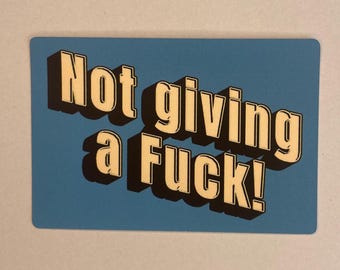 May include: A rectangular card with the phrase "Not giving a Fuck!" in bold, off-white letters with a black shadow effect. The text is centered on a light blue background.