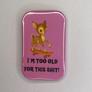 Könnte beinhalten: Ein rechteckiger, rosa Button mit abgerundeten Ecken. Er zeigt ein Cartoon-Reh auf einem Skateboard. Der Text "I'M TOO OLD FOR THIS SHIT!" ist darunter in Schwarz gedruckt.