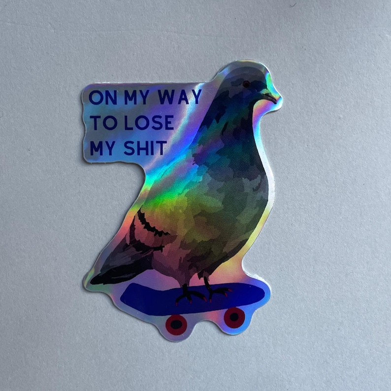 May include: A holographic sticker featuring a pigeon standing on a blue skateboard. The text ON MY WAY TO LOSE MY SHIT is printed in dark blue letters on the left side. The sticker has a reflective, iridescent finish that shifts colors.