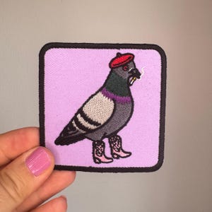 Könnte beinhalten: Ein quadratischer gestickter Aufnäher mit schwarzem Rand auf hellviolettem Hintergrund. Das Design zeigt eine graue Taube mit rotem Barett und rosa Cowboystiefeln, die eine kleine Zigarette im Schnabel hält, ausgeführt in detaillierter Stickerei.
