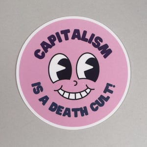 Könnte beinhalten: Ein runder Aufkleber mit rosa Hintergrund und weißem Rand. Der Aufkleber zeigt ein Cartoon-Gesicht mit großen Augen und einem lächelnden Mund. Der Text "CAPITALISM IS A DEATH CULT!" ist in lila Schrift um das Gesicht geschrieben.