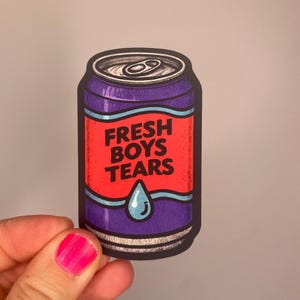 Könnte beinhalten: Aufkleber in Form einer lila Getränkedose mit rotem Etikett. Auf dem Etikett steht in weißer Schrift "FRESH BOYS TEARS". Am unteren Rand befindet sich eine blaue Tränen-Grafik.