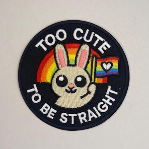 Może przedstawiać: Okrągła czarna naszywka z napisem "TOO CUTE TO BE STRAIGHT". Naszywka przedstawia kreskówkowego królika trzymającego tęczową flagę z sercem i tęczą za królikiem.