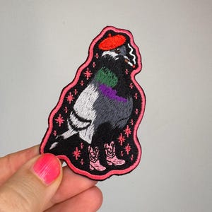 Könnte beinhalten: Ein gestickter Aufnäher mit einer Taube, die ein rotes Barett und rosa Cowboystiefel trägt. Die Taube hat grüne und violette Halsfedern, einen weißen Flügel und hält eine Zigarette. Der Hintergrund ist schwarz mit rosa Sternen und einem rosa Rand.