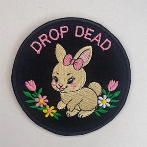 Könnte beinhalten: Runder, gestickter Aufnäher mit schwarzem Hintergrund. Er zeigt ein beiges Häschen mit rosa Schleife, umgeben von Blumen. Der Text "DROP DEAD" ist in rosa Buchstaben über dem Hasen angeordnet.