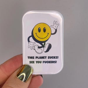 Könnte beinhalten: Ein weißer, rechteckiger Ansteck-Pin mit einem gelben Smiley-Gesicht. Die Figur hat schwarze Augen, einen schwarzen Umriss und winkt. Der Pin hat den Text "THIS PLANET SUCKS! SEE YOU FUCKERS!" in Schwarz.