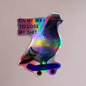 May include: A holographic sticker featuring a pigeon riding a skateboard. The bird is depicted in shades of dark gray and purple. The text ON MY WAY TO LOSE MY SHIT is printed in dark blue capital letters to the left of the pigeon's head.