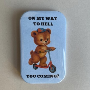 Könnte beinhalten: Rechteckiger Ansteck-Pin mit hellblauem Hintergrund, der einen braunen Teddybären auf einem roten Roller zeigt. Der Text "ON MY WAY TO HELL" steht über dem Bären und "YOU COMING?" darunter.