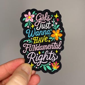 May include: Embroidered patch with the text "Girls Just Wanna Have Fundamental Rights" in white, pink, and yellow lettering. The patch is black with floral accents in pink, orange, and green.