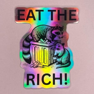 May include: Holographic sticker featuring two raccoons and a trash can. The text "EAT THE RICH!" is displayed above and below the illustration. The raccoons are depicted in black and white with colorful accents.