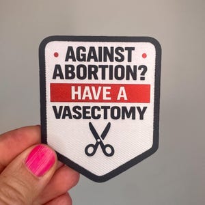 Könnte beinhalten: Ein weiß-schwarzer Aufnäher mit dem Text "AGAINST ABORTION? HAVE A VASECTOMY" in fetten, schwarzen Buchstaben. Ein roter horizontaler Balken trennt die beiden Sätze. Am unteren Rand befindet sich eine Schere. Der Aufnäher wird von einer Hand mit pinkfarbenem Nagellack gehalten.