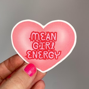 Könnte beinhalten: Ein rosa herzförmiger Aufkleber mit den Worten "MEAN GIRL ENERGY" in roter und rosa Schrift. Der Aufkleber hat einen weißen Rand. Der Aufkleber wird von einer Hand gehalten.