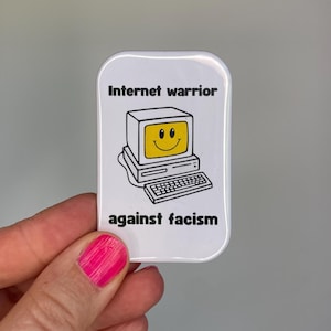 Könnte beinhalten: Ein weißer, rechteckiger Knopf mit abgerundeten Ecken. Der Knopf zeigt einen Cartoon-Computer mit einem lächelnden Gesicht auf dem Bildschirm. Der Text auf dem Knopf lautet "Internet warrior" über dem Computer und "against facism" darunter.